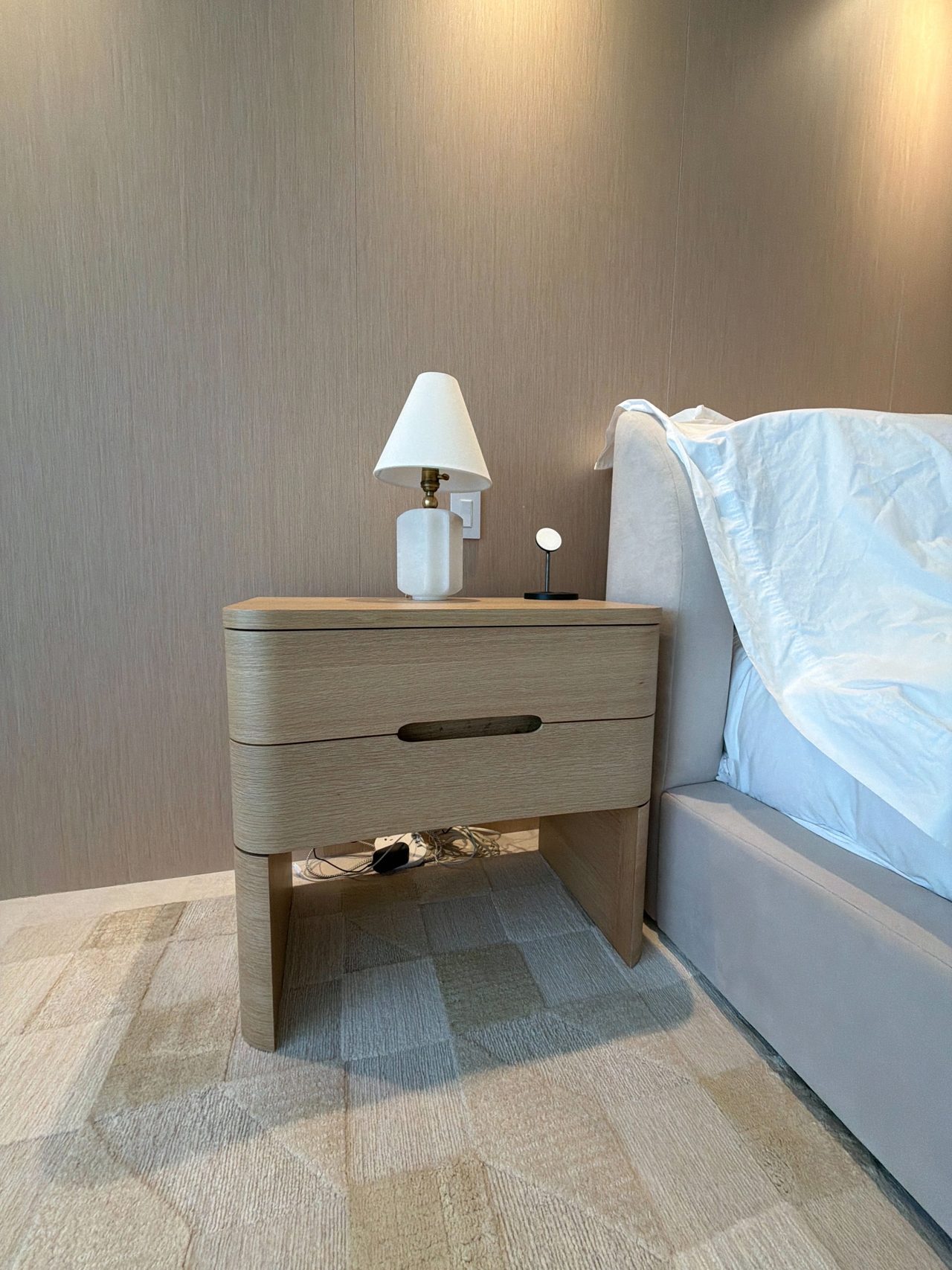 Close view of a rounded custom-stained wood veneer nightstand with two finger-pull drawers, a table lamp, and a downlit veneer wall