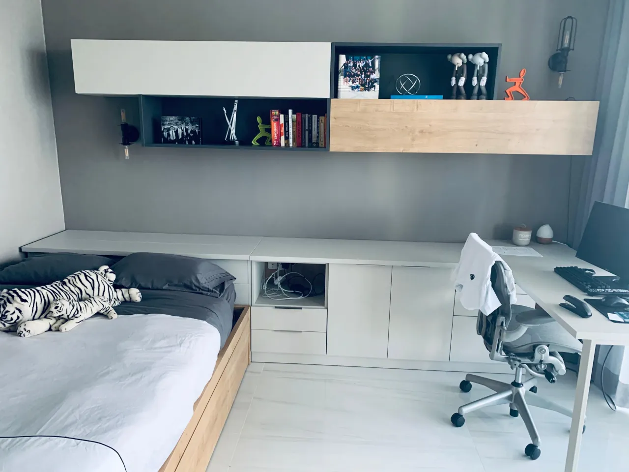 Teen room with gray wall-mounted floating shelves and dark box shelving over a built-in desk with monitor, tiger plush toys on bed