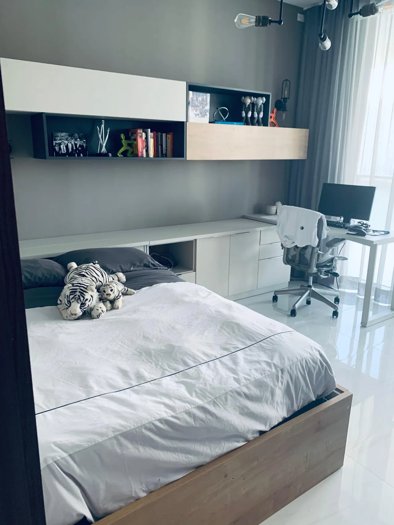 Teen bedroom wall unit with gray and white built-in storage cabinets, floating shelves, integrated desk with monitor, tiger plush toys on the bed