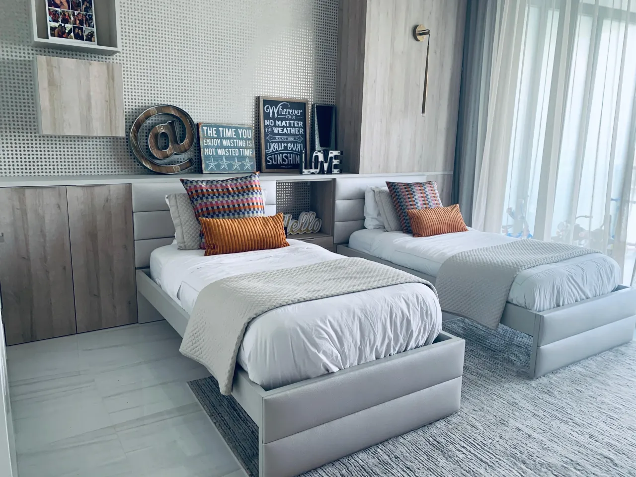 Twin beds with a built-in upholstered headboard unit and open cubby, textured dot-pattern wall, wood-grain cabinet doors, window with sheer curtains
