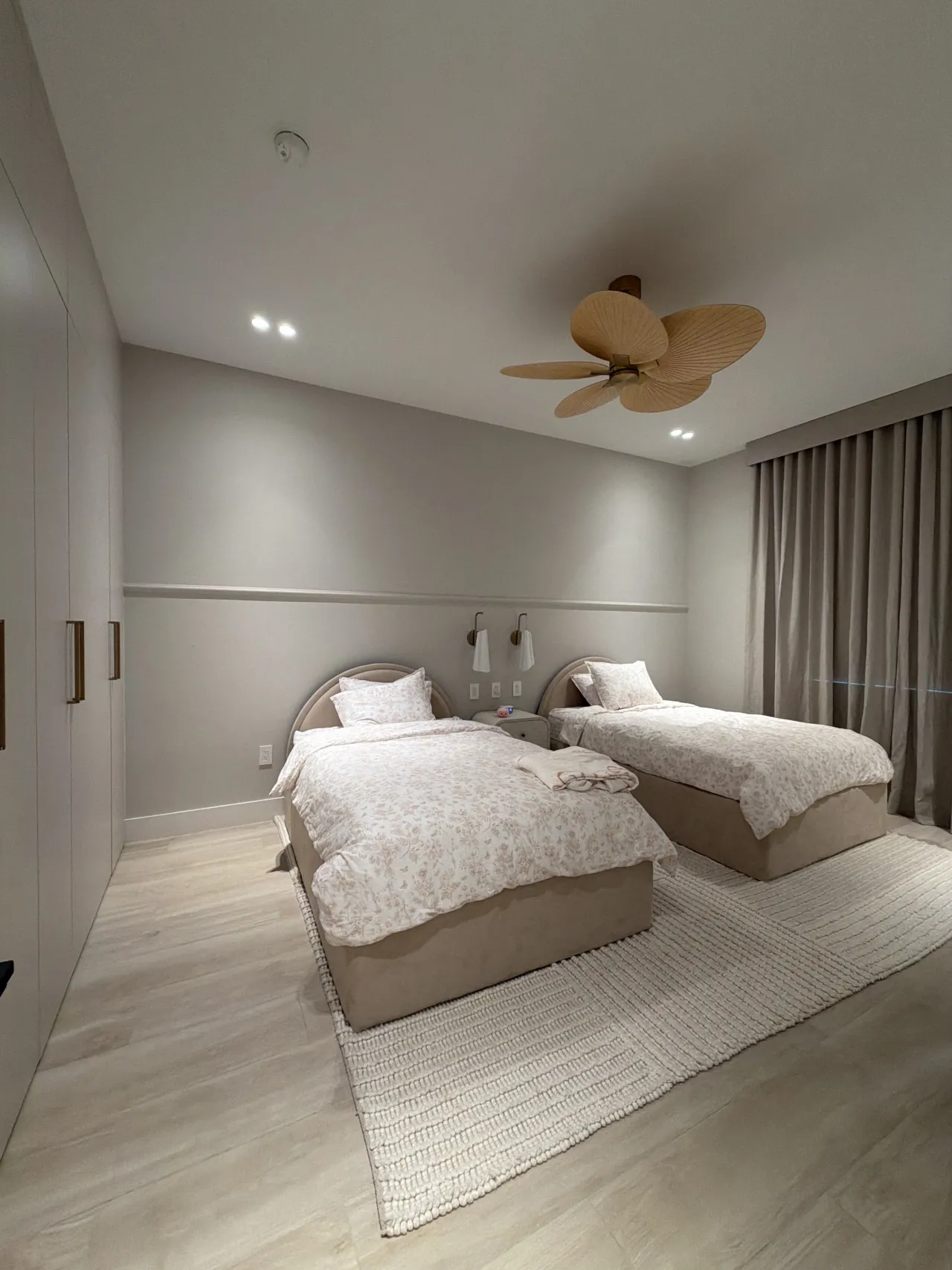 Twin beds with pale floral bedding and beige upholstered bases, woven palm-blade ceiling fan, greige walls with a rail, wall sconces, textured rug