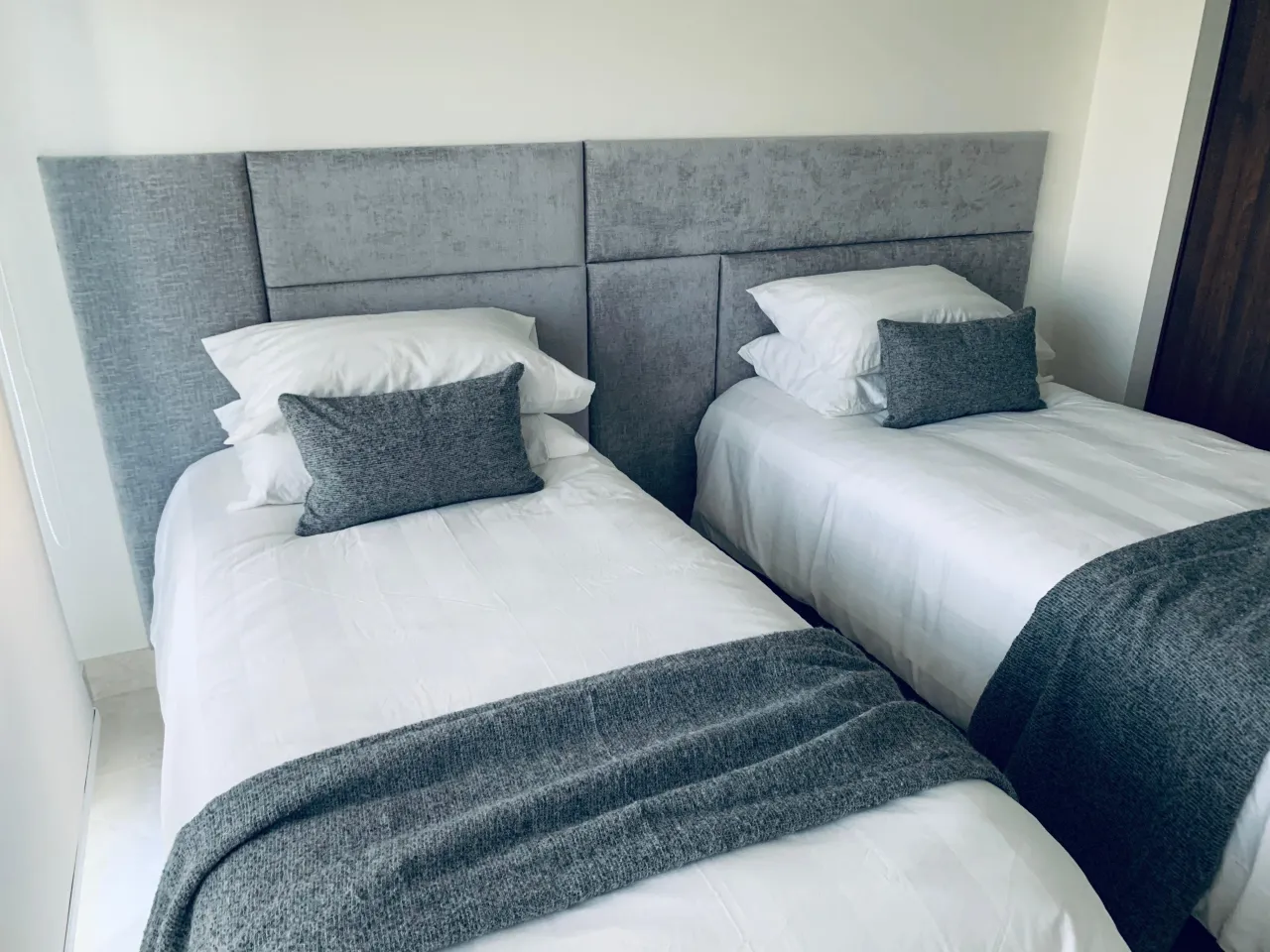 Two twin beds with white bedding and gray knit throws against a tall gray upholstered headboard wall divided into rectangular panels