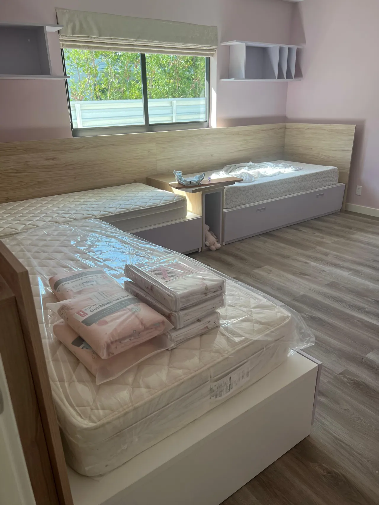 Unfinished pink-walled girls room with wood built-in twin bed frames and headboard surround, mattresses still wrapped in plastic