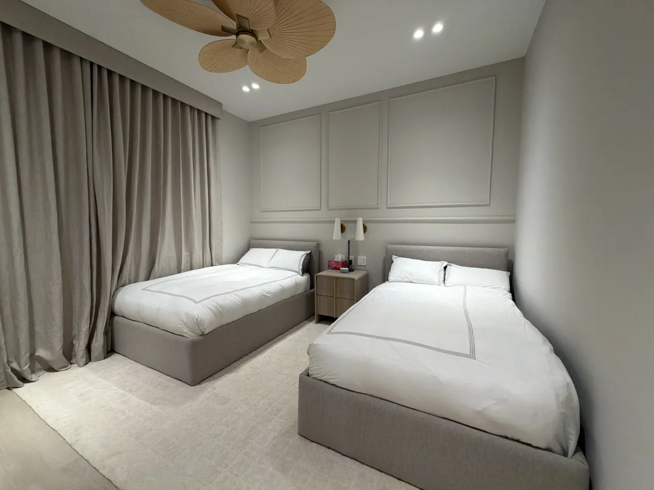 Two upholstered twin beds with white bedding against a greige molded panel wall, wood-grain nightstand, floor-length drapes, palm-blade fan