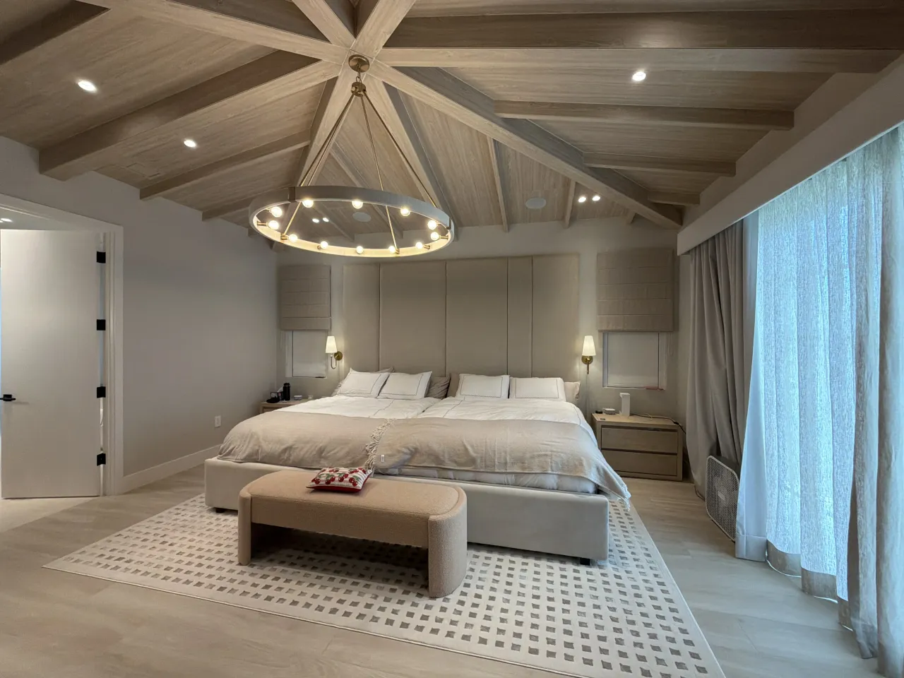 Bedroom with a vaulted wood-grain melamine-clad ceiling and beams, round metal ring chandelier, tall beige upholstered headboard, bench at bed foot