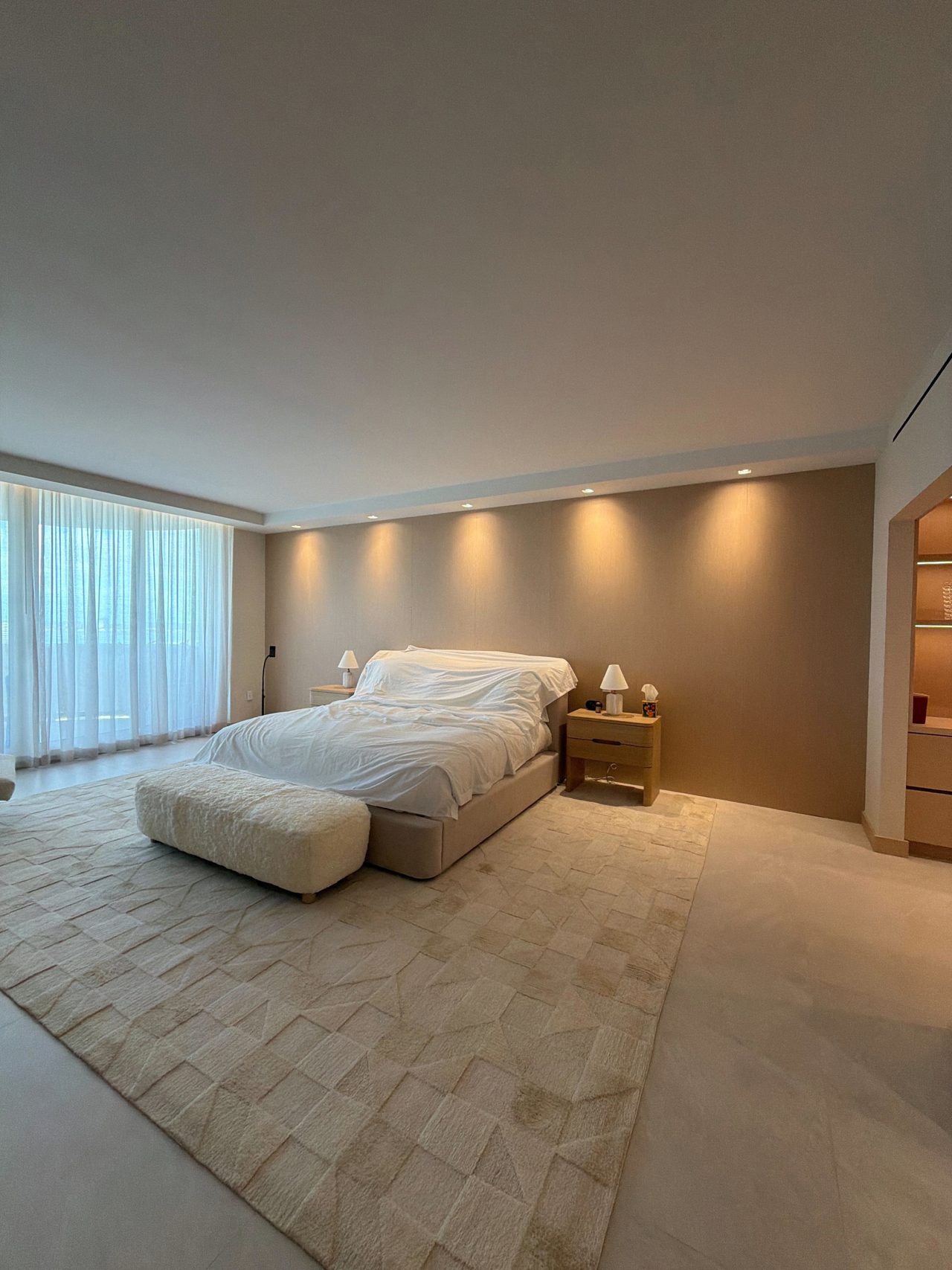 Bed against a full-height veneer headboard wall lit by a row of recessed downlights, with a veneer nightstand and an upholstered bench