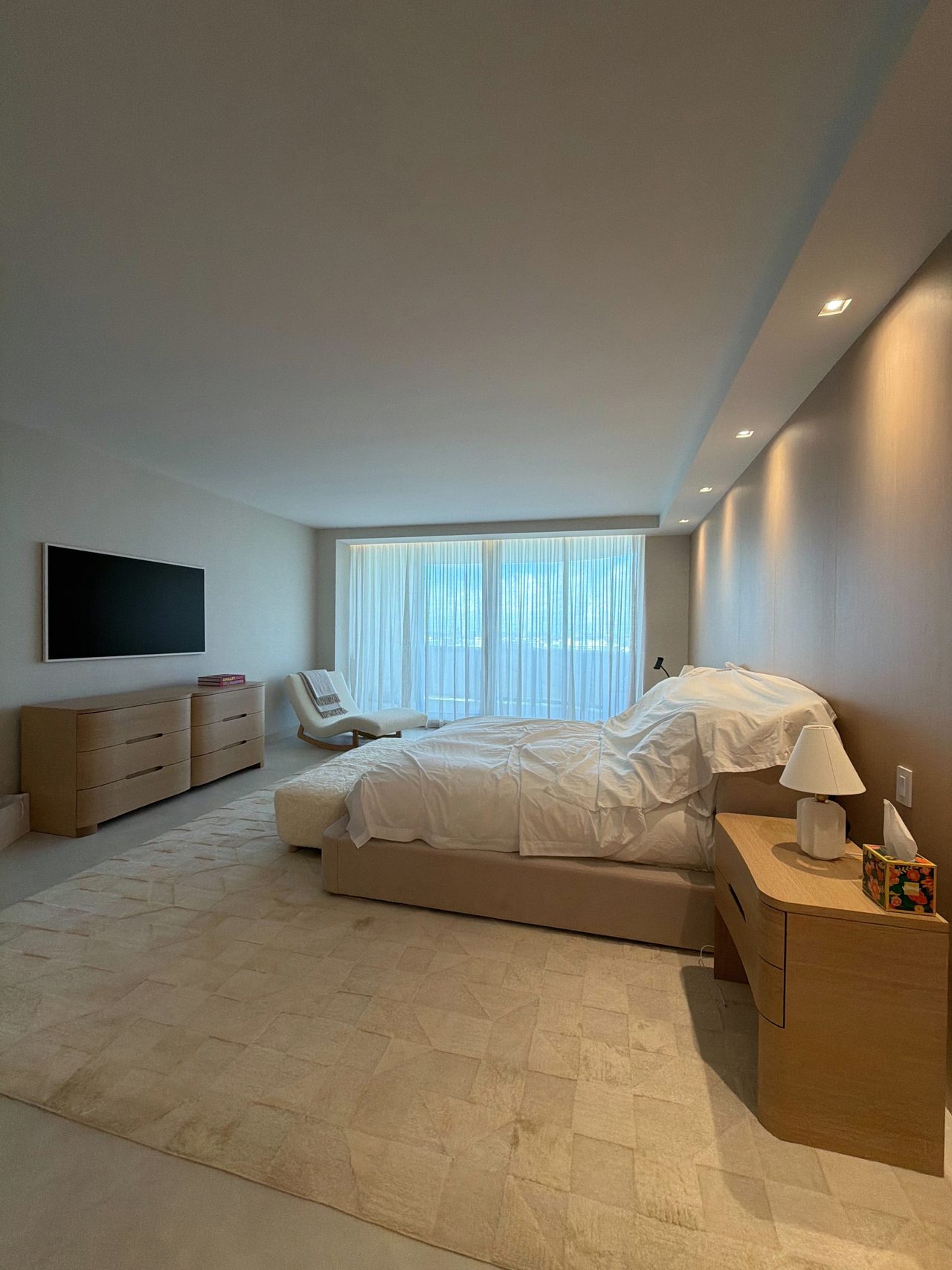 Primary bedroom with a veneer headboard wall washed by recessed downlights, a low veneer dresser, a nightstand, and a wall-mounted TV
