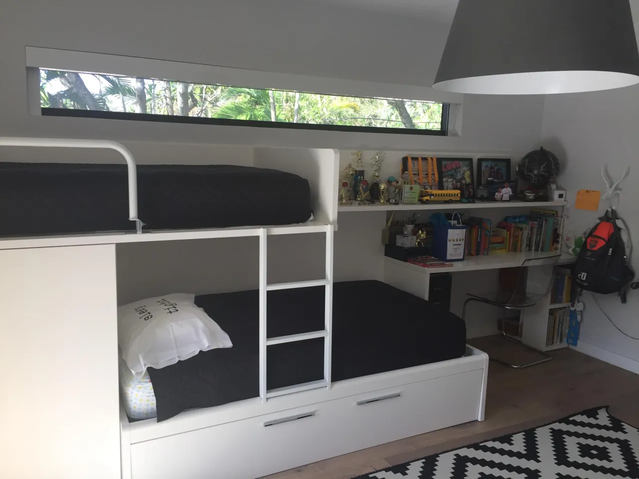 White built-in bunk bed unit with black mattresses, a ladder, trundle drawer below, and a long window strip above
