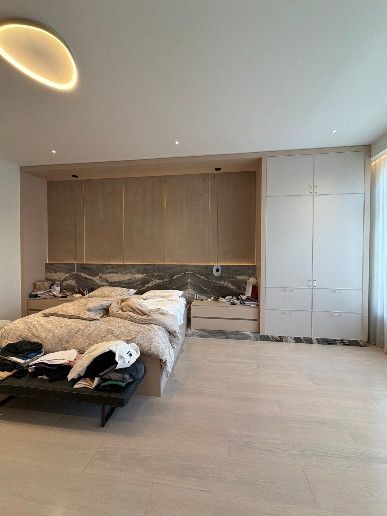 Bedroom with a wood-grain overhead cabinet run over the bed, against a dark veined stone wall, tall white closet unit at right, clothes on a black bench