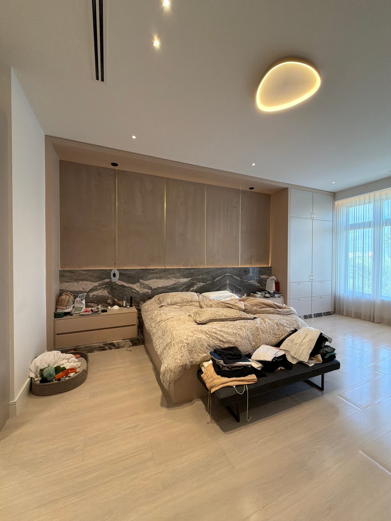 Bedroom with wood-grain overhead cabinets above the bed, set against a dark veined stone wall, tall white closet doors, black bench, light tile floor