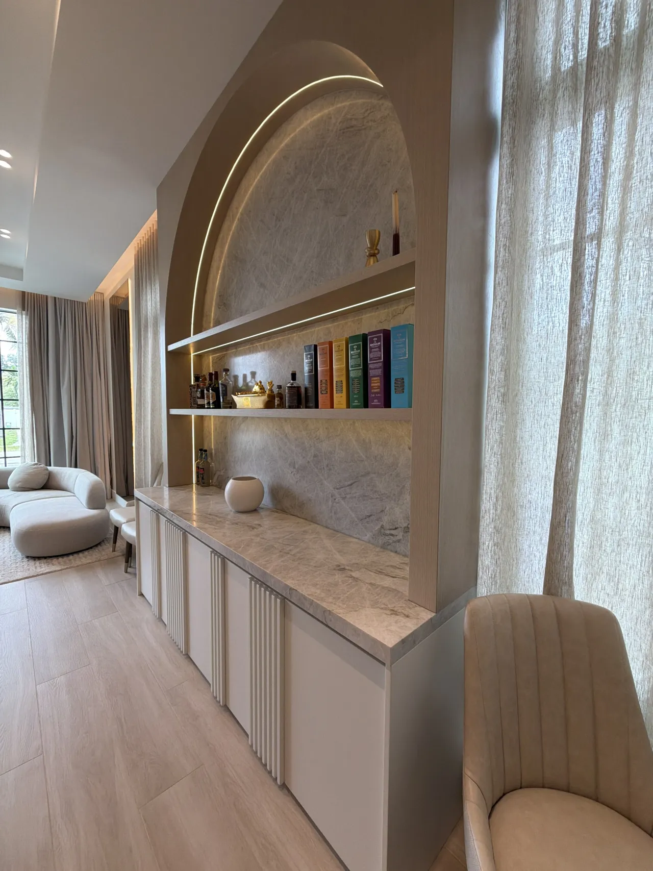 Angled view of an arched LED-lit niche with veined stone back and shelves, over a white credenza with fluted fronts, sheer curtain at right
