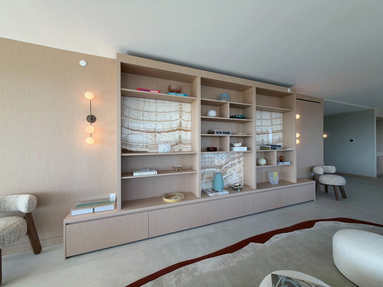 Angled view of a full-height custom-stained wood veneer built-in with open shelving, backlit veined-stone niches, and a floating credenza base