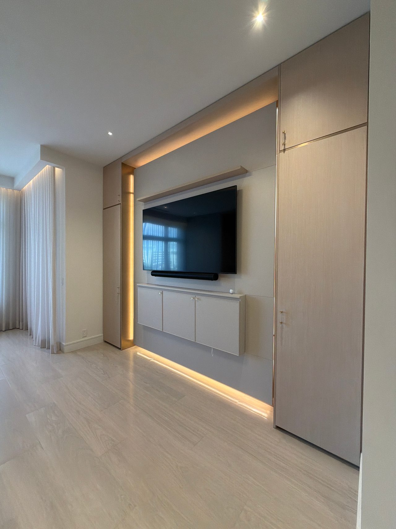 Angled view of a recessed TV wall with backlit trim, a white wall-hung cabinet below the screen and a tall wood-grain melamine cabinet beside it