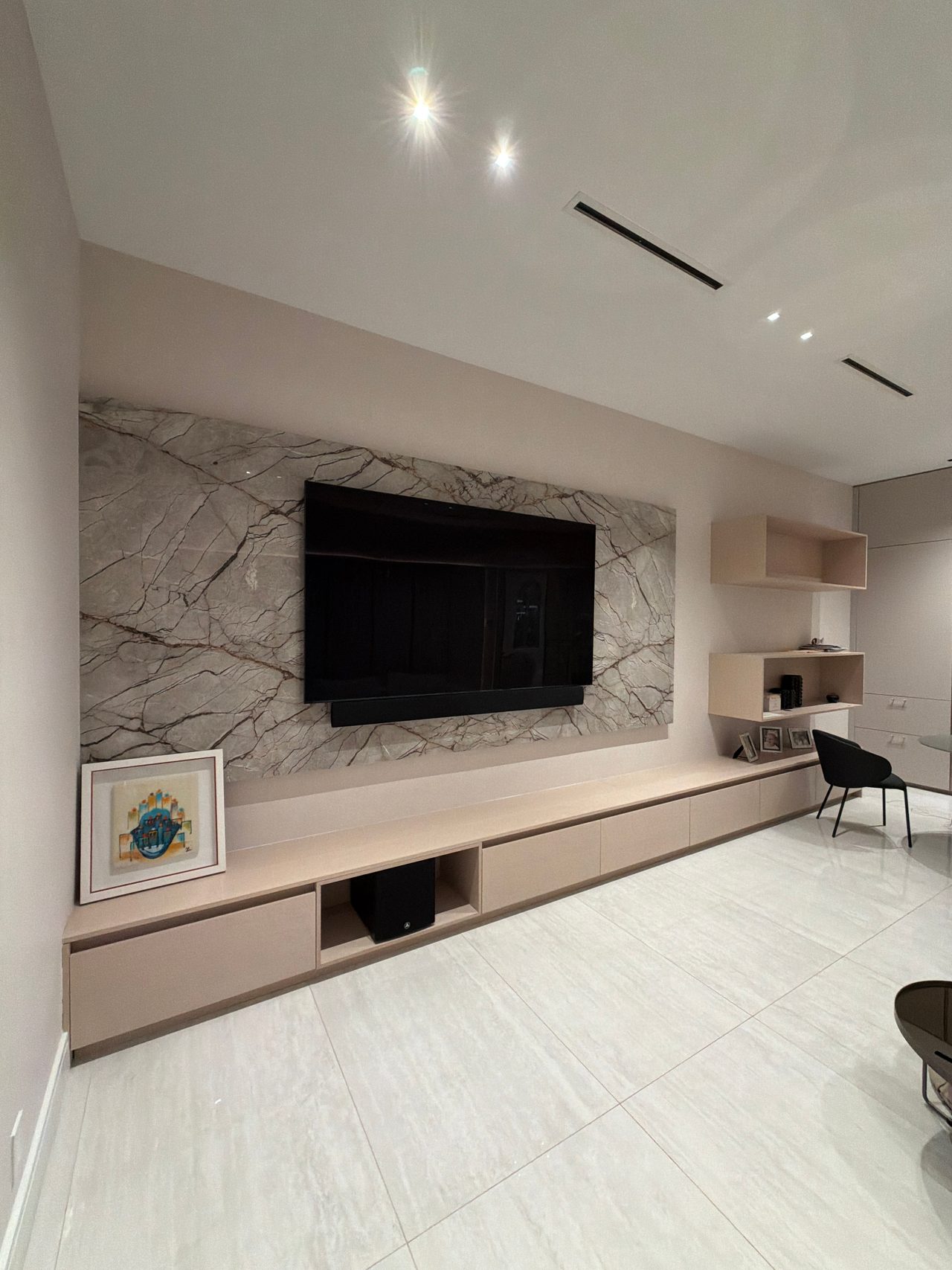 Angled view of a veined stone wall with a mounted TV above a long low wood-grain credenza, framed art leaning at the end and open shelf boxes right