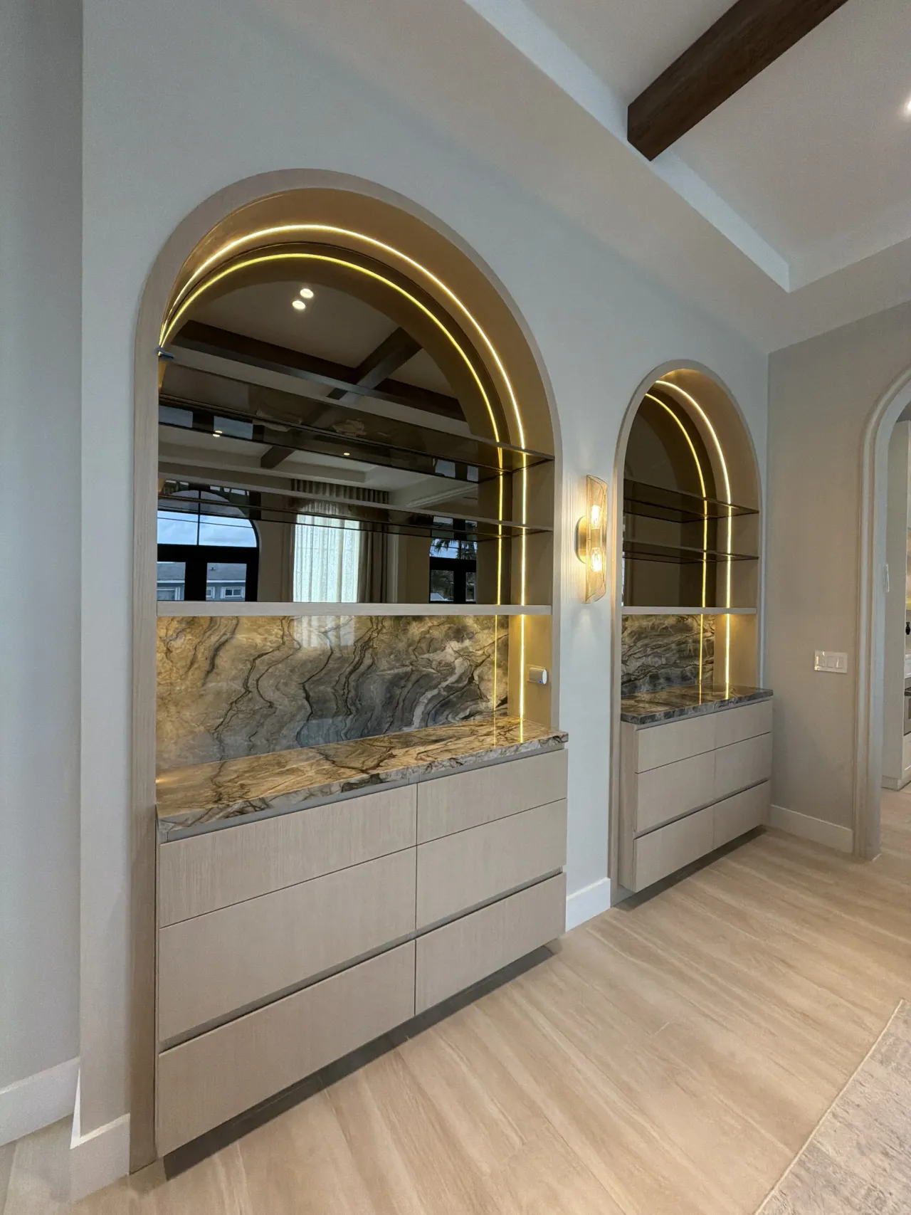 Two arched display niches with gold-tone LED trim, mirrored and veined stone backs, and glass shelves, over light wood-grain drawer bases