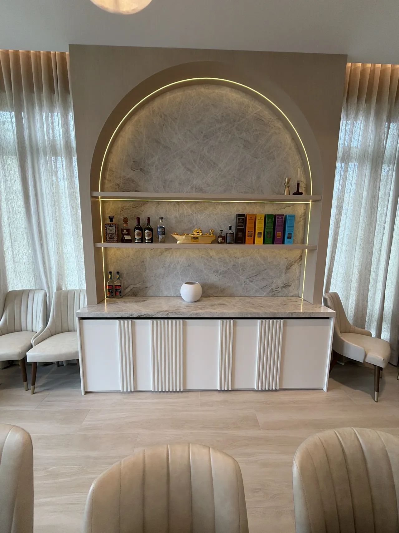 Arched niche with LED trim, veined light stone back and two floating shelves holding bottles, over a white credenza with fluted fronts and stone top