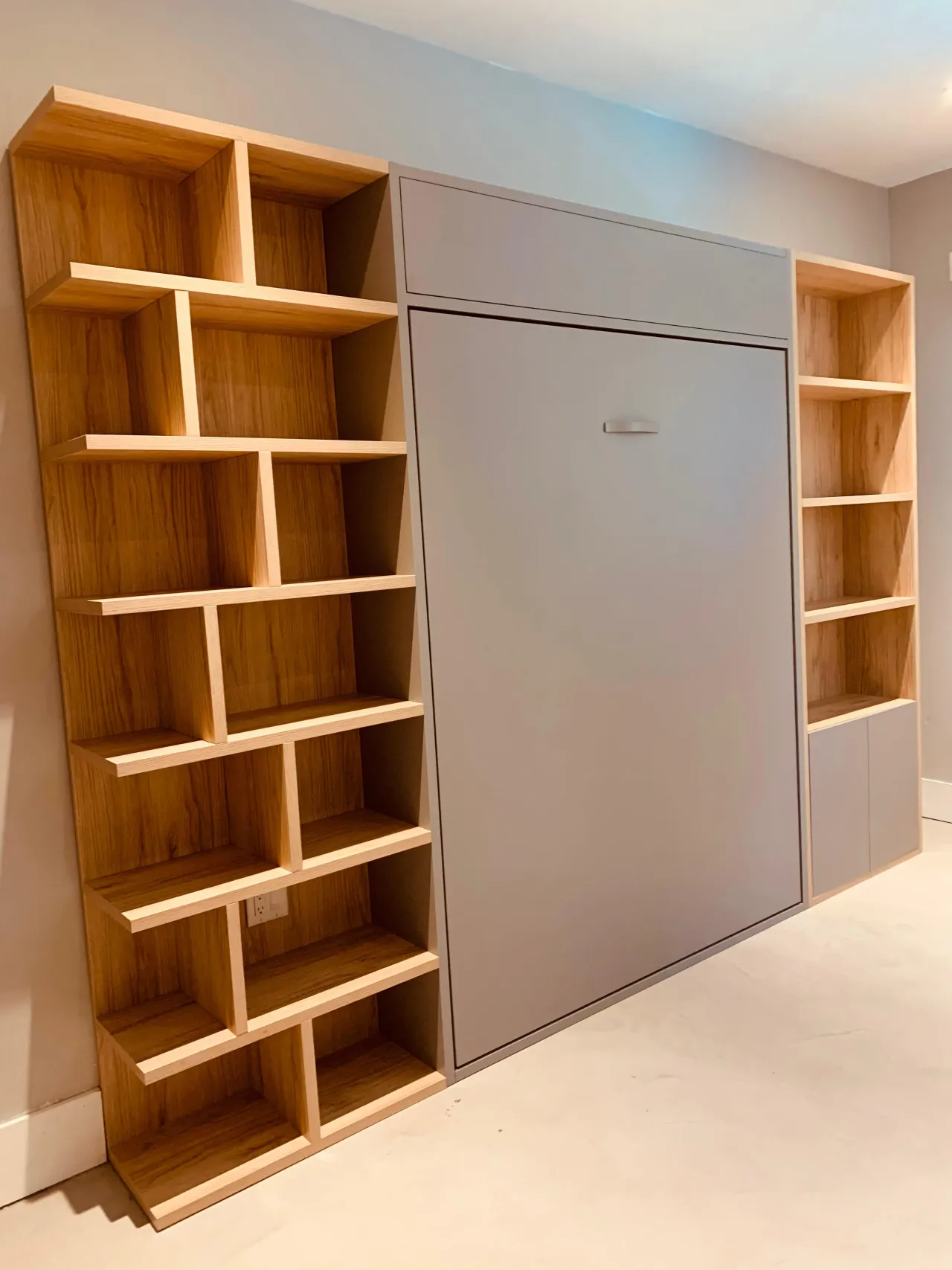 Asymmetric light wood bookshelf with angled open cubbies flanking a gray murphy bed panel