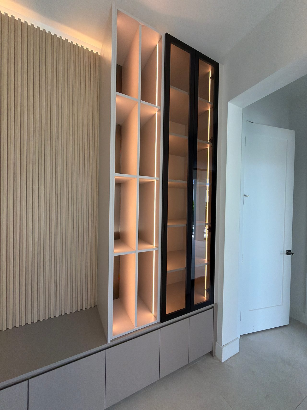 Backlit white grid shelving column next to a tall black-framed glass cabinet, over low gray cabinet fronts, wood-grain slat wall at left