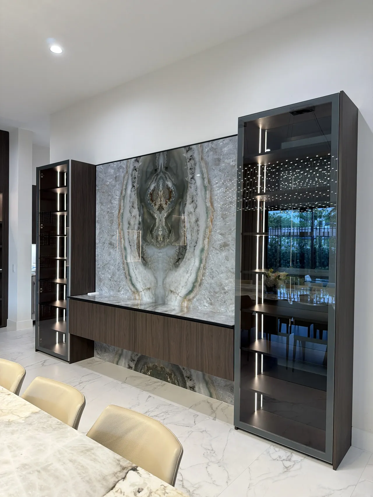 Bar cabinet with a large gray-green stone slab backsplash, flanked by tall glass display towers with LED strip lighting
