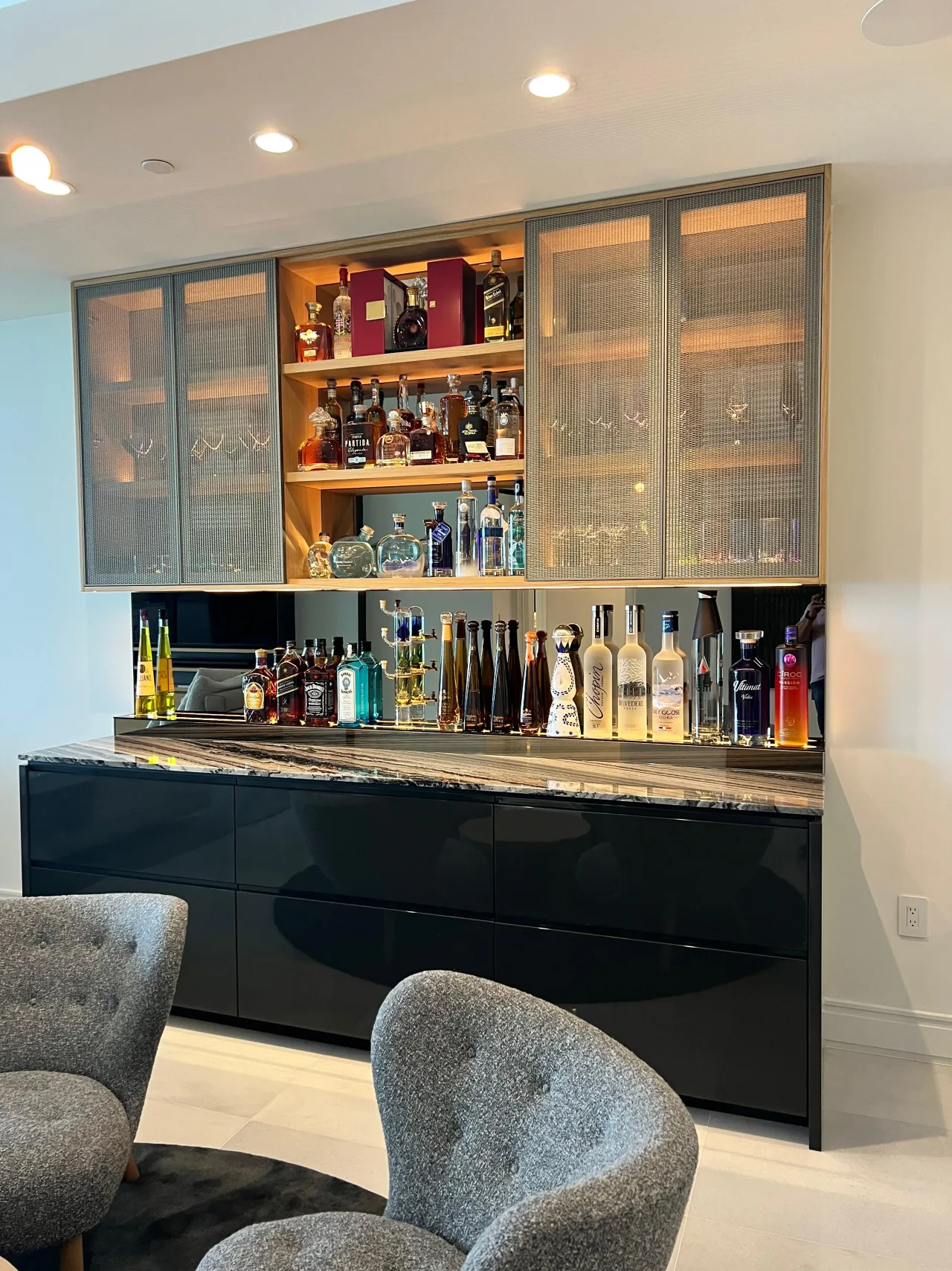 Bar wall with mesh-front glass upper doors, lit open shelves of bottles and glassware, mirrored backsplash, veined stone counter, black gloss base