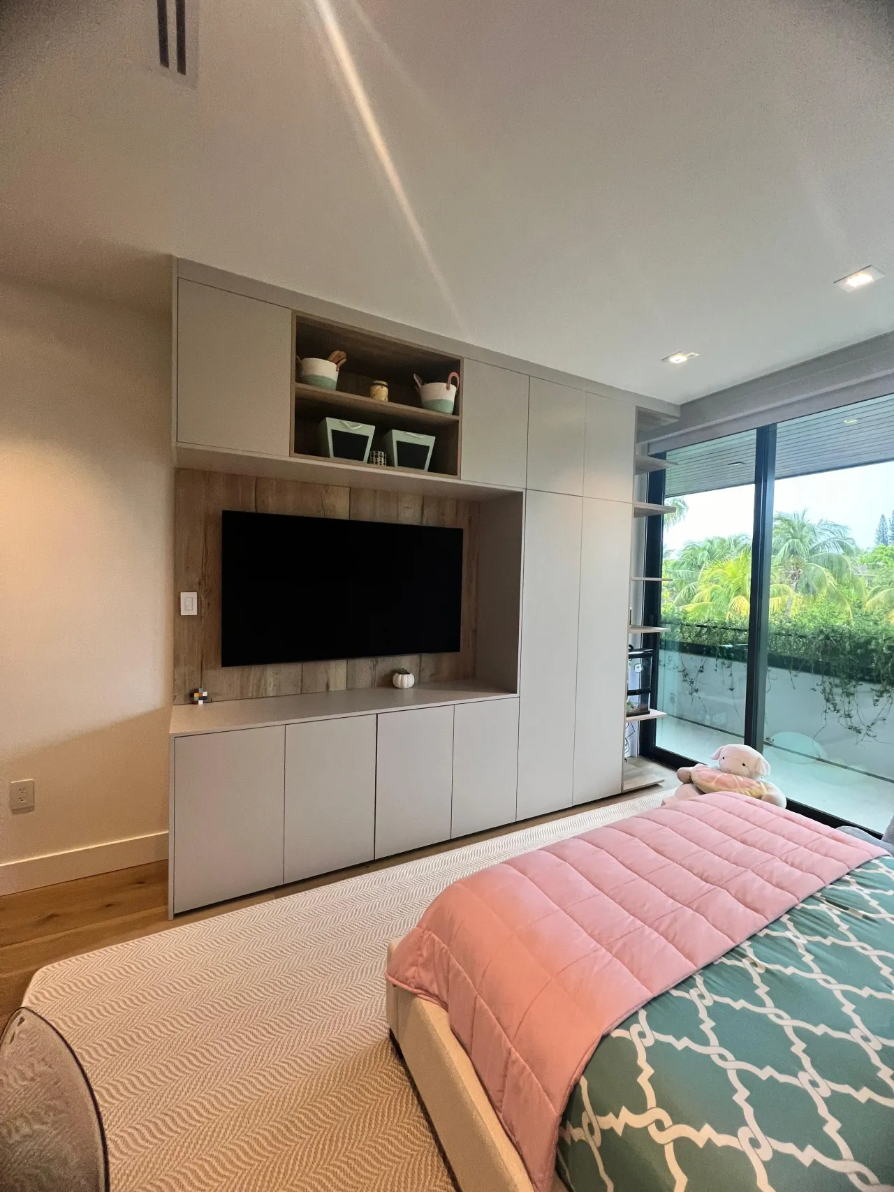 Bedroom built-in TV wall unit with a light wood accent panel behind the screen, flanked by white cabinets, glass door to a palm-view balcony