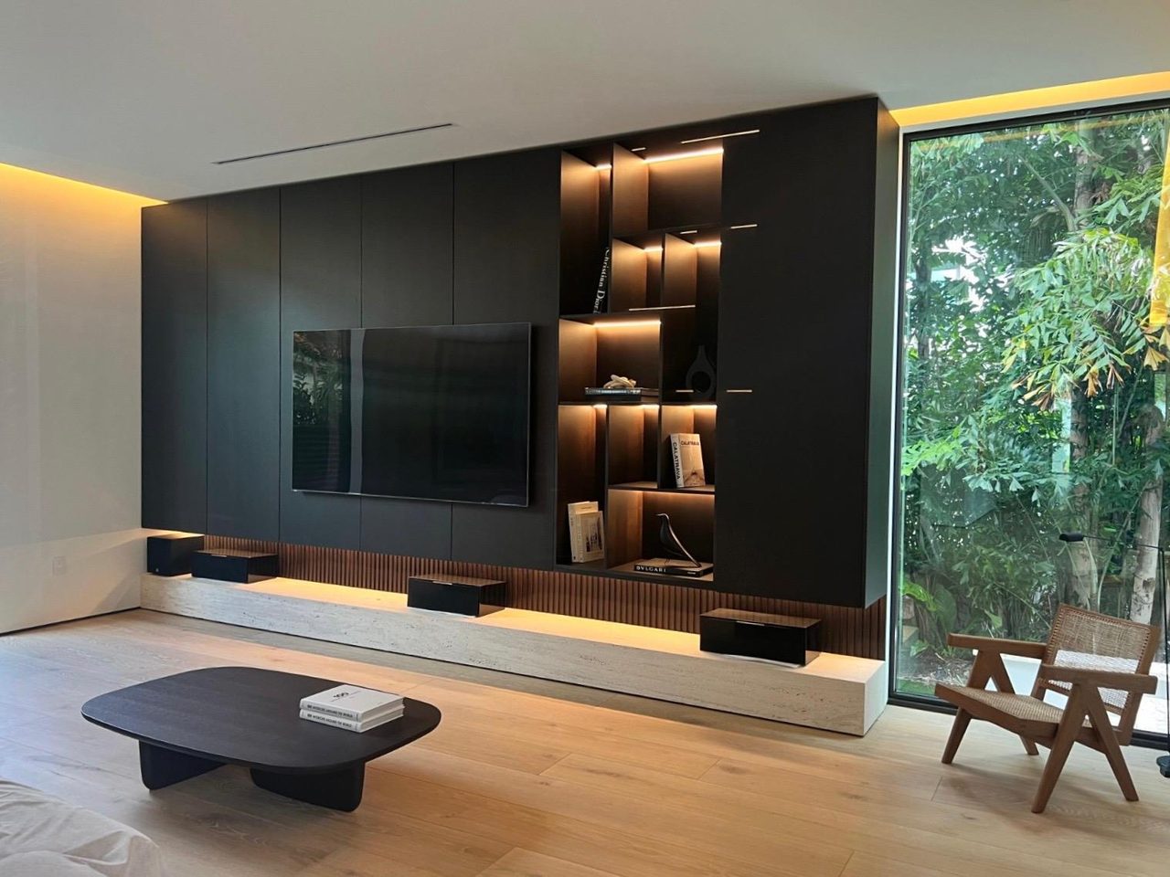 Black built-in wall unit with a mounted TV and backlit angled display cubbies above a fluted band, tall window beside it