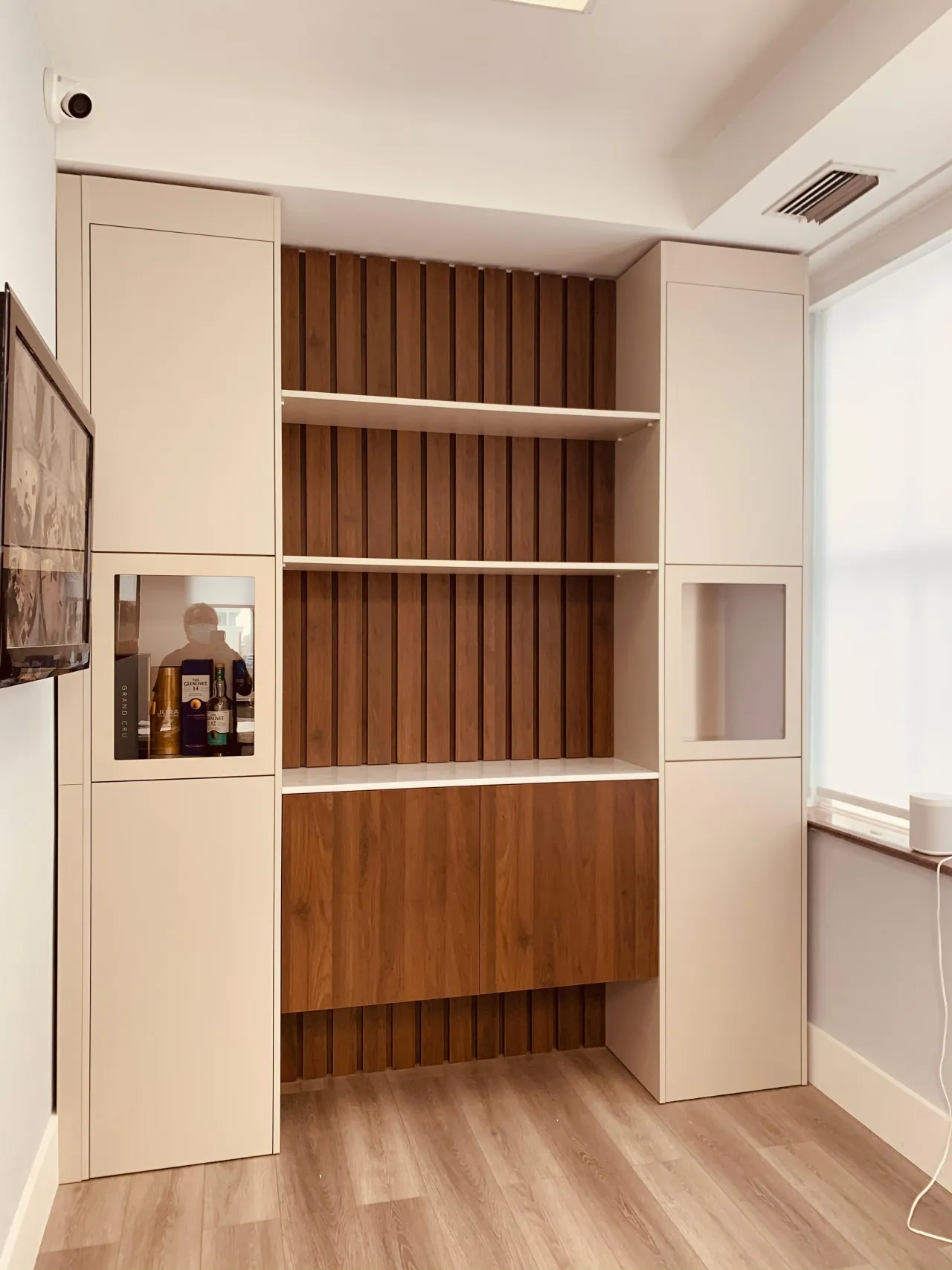 Built-in bar niche with vertical wood slat backing, open shelves, and bottle display, flanked by beige cabinet towers with glass insets