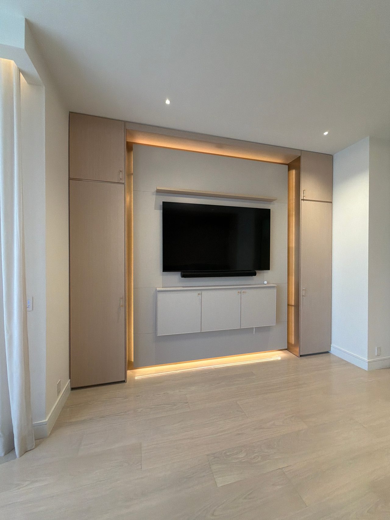 Built-in TV niche with backlit trim, wood-grain melamine tall cabinets on each side and a white wall-hung cabinet below the mounted screen