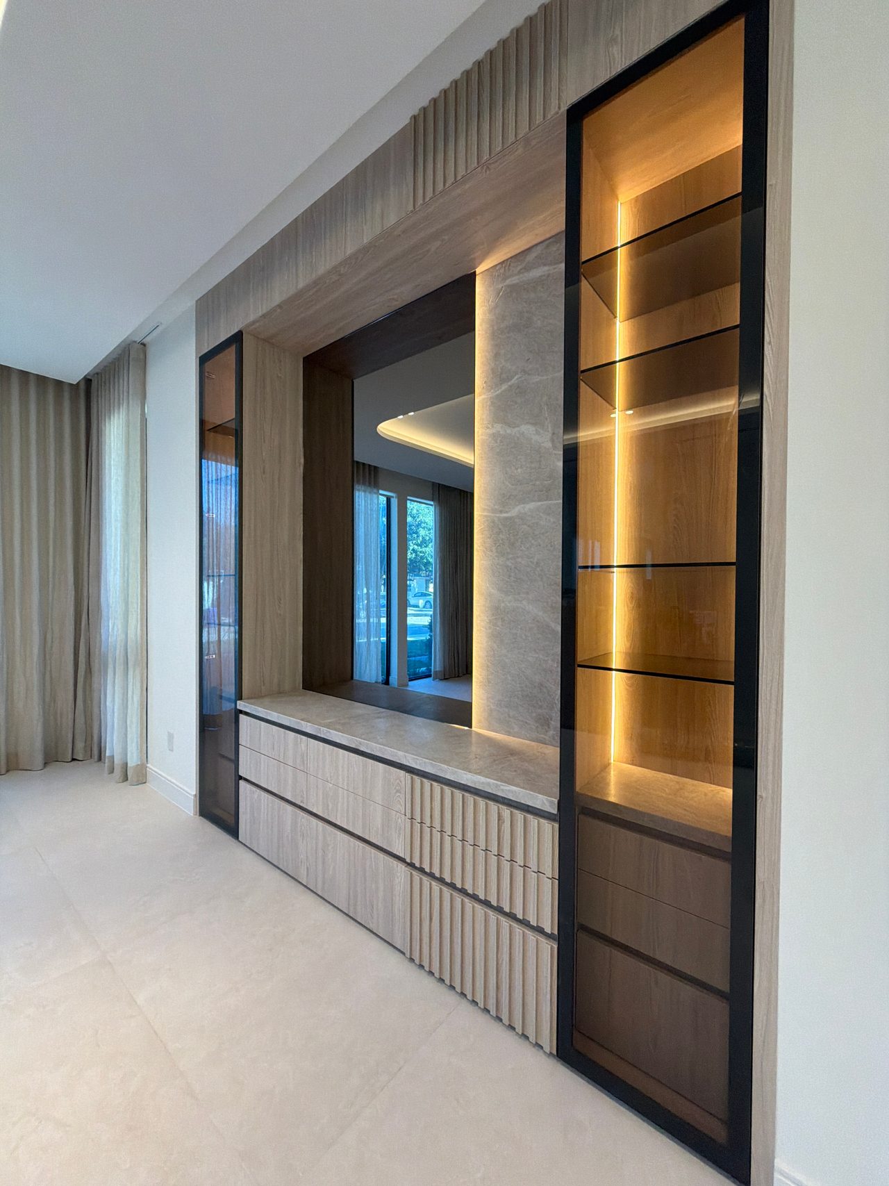 Built-in wall unit with a black-framed glass display cabinet, backlit shelves, wood-grain fluted drawers, a stone-look wall and a mirrored recess