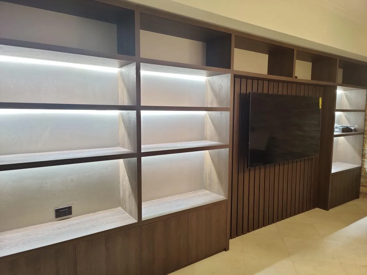 Built-in wall unit in dark wood-grain with LED-lit open shelving, lower cabinet doors, and a wall-mounted TV on a vertical slat panel section