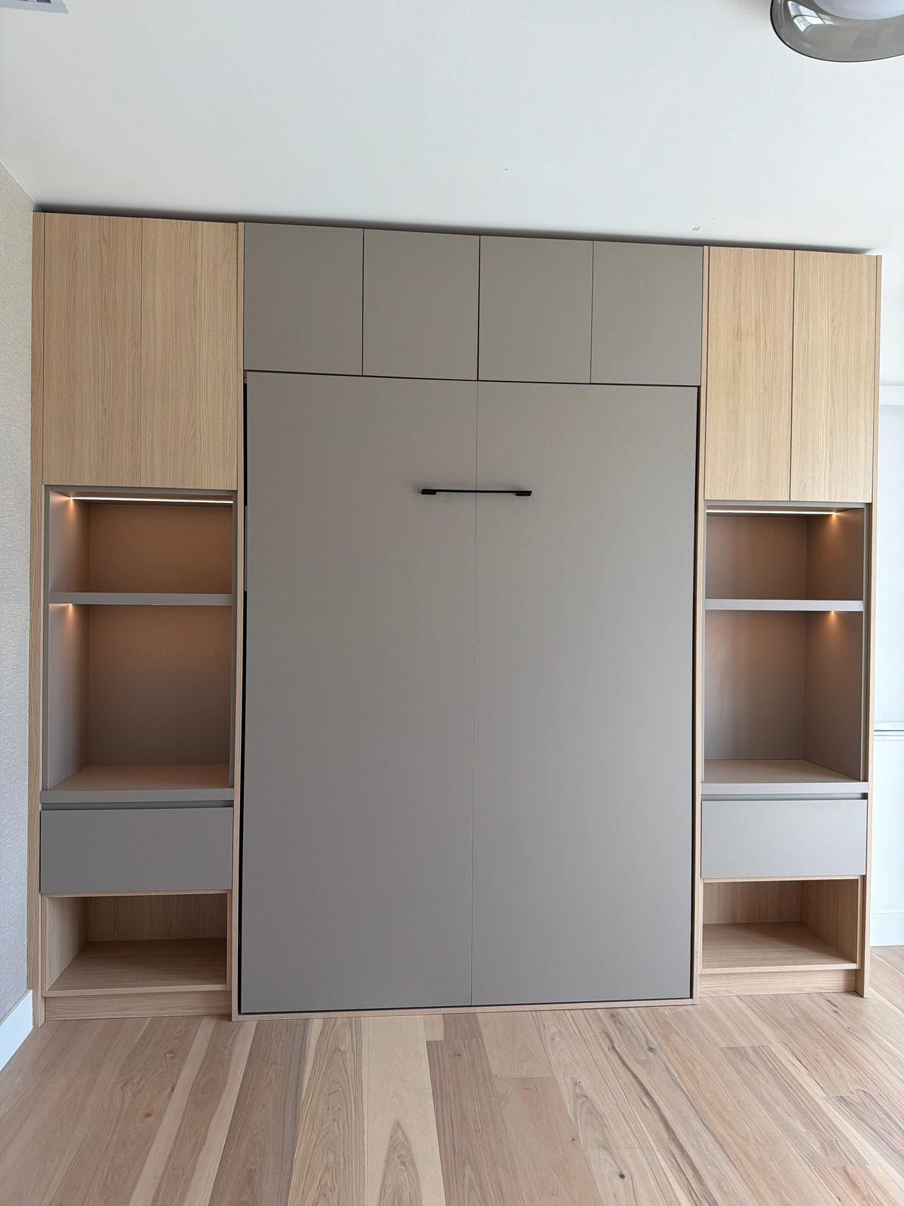 Built-in wall unit with light wood-grain and gray cabinetry framing a large gray fold-down panel, flanked by lit open shelves and drawers