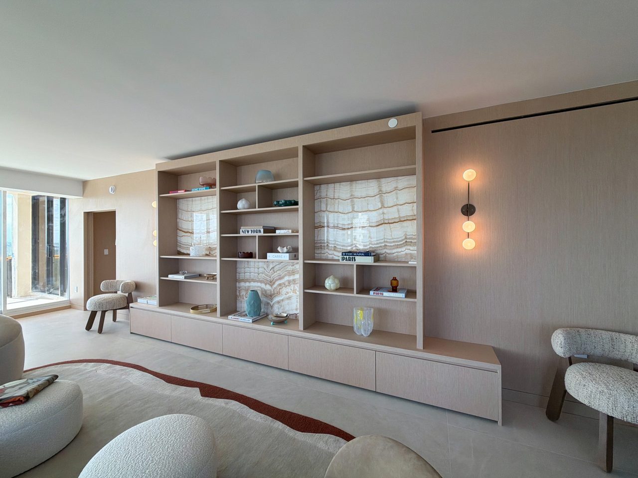Custom-stained wood veneer built-in shelving wall with three backlit veined-stone niches and a low drawer base, flanked by three-globe wall sconces