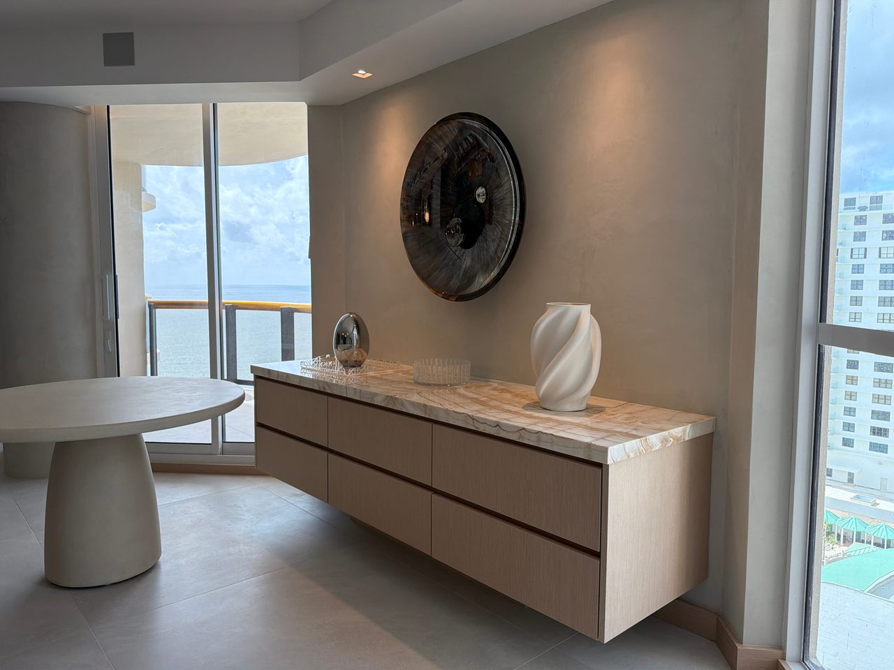 Floating six-drawer credenza in custom-stained wood veneer with a veined stone top, beside a round dining table and balcony doors