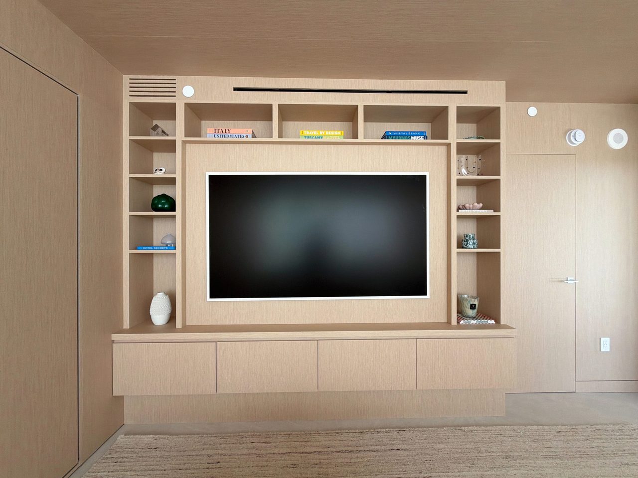 Front view of a custom-stained wood veneer TV built-in: recessed white-framed screen, open cubby shelving, and a four-drawer floating base