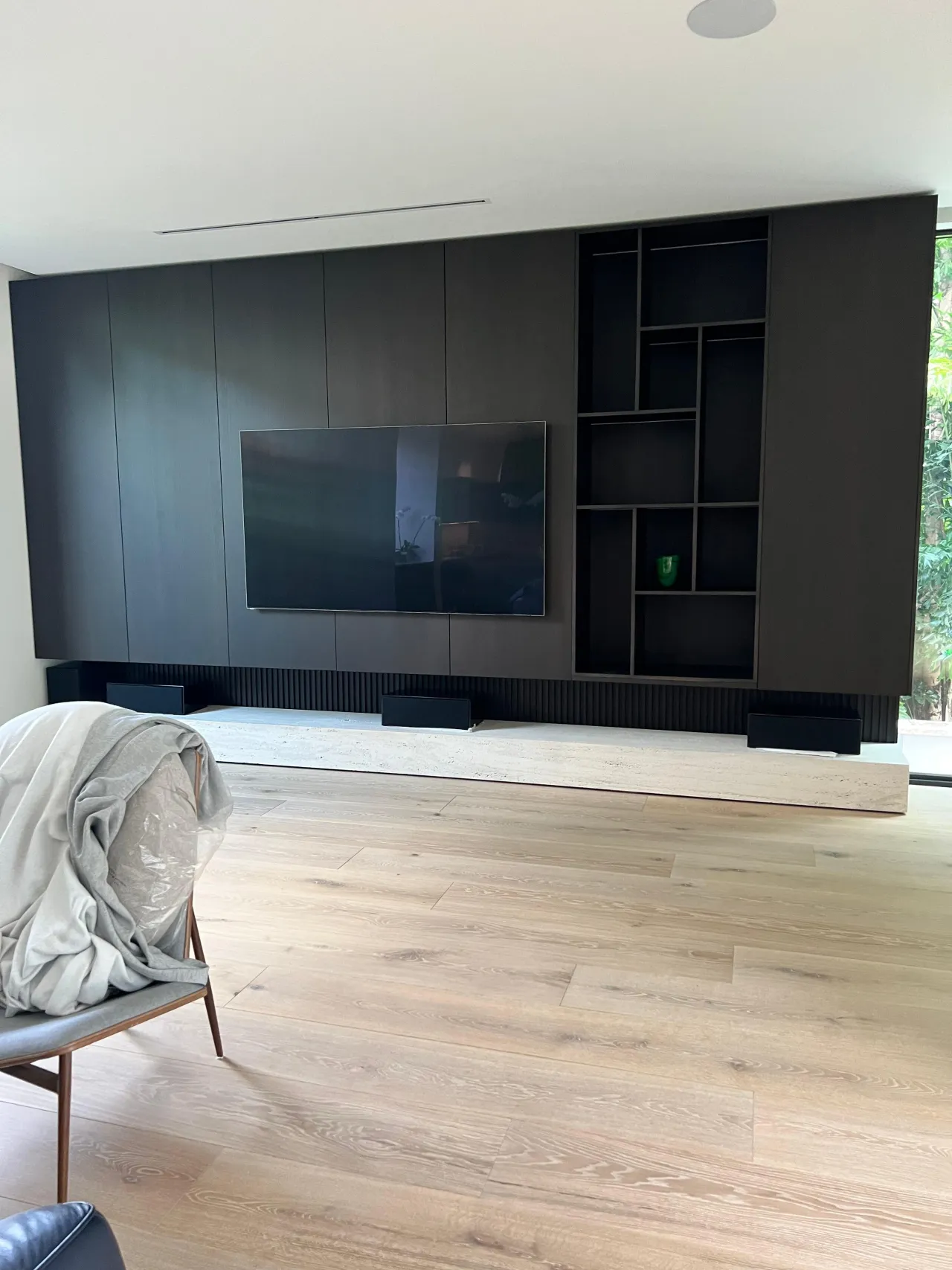 Dark charcoal flat-panel media wall with wall-mounted TV and an open asymmetric geometric shelving cutout, on a light stone-look base
