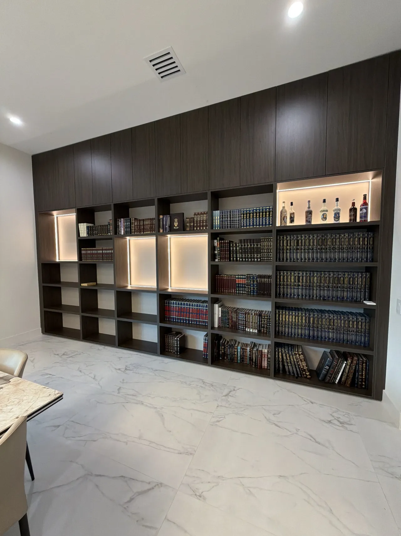 Dark espresso library wall with backlit niches, now showing a bar area with bottles on lit shelves