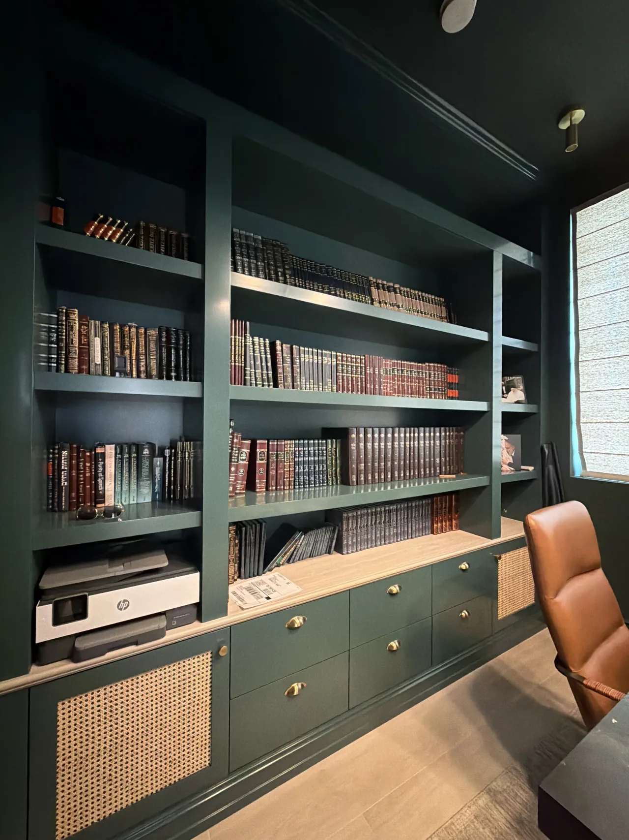 Dark green lacquered library shelving with rows of books, a printer nook, and lower cabinets with cane/rattan panel fronts and gold-tone pulls