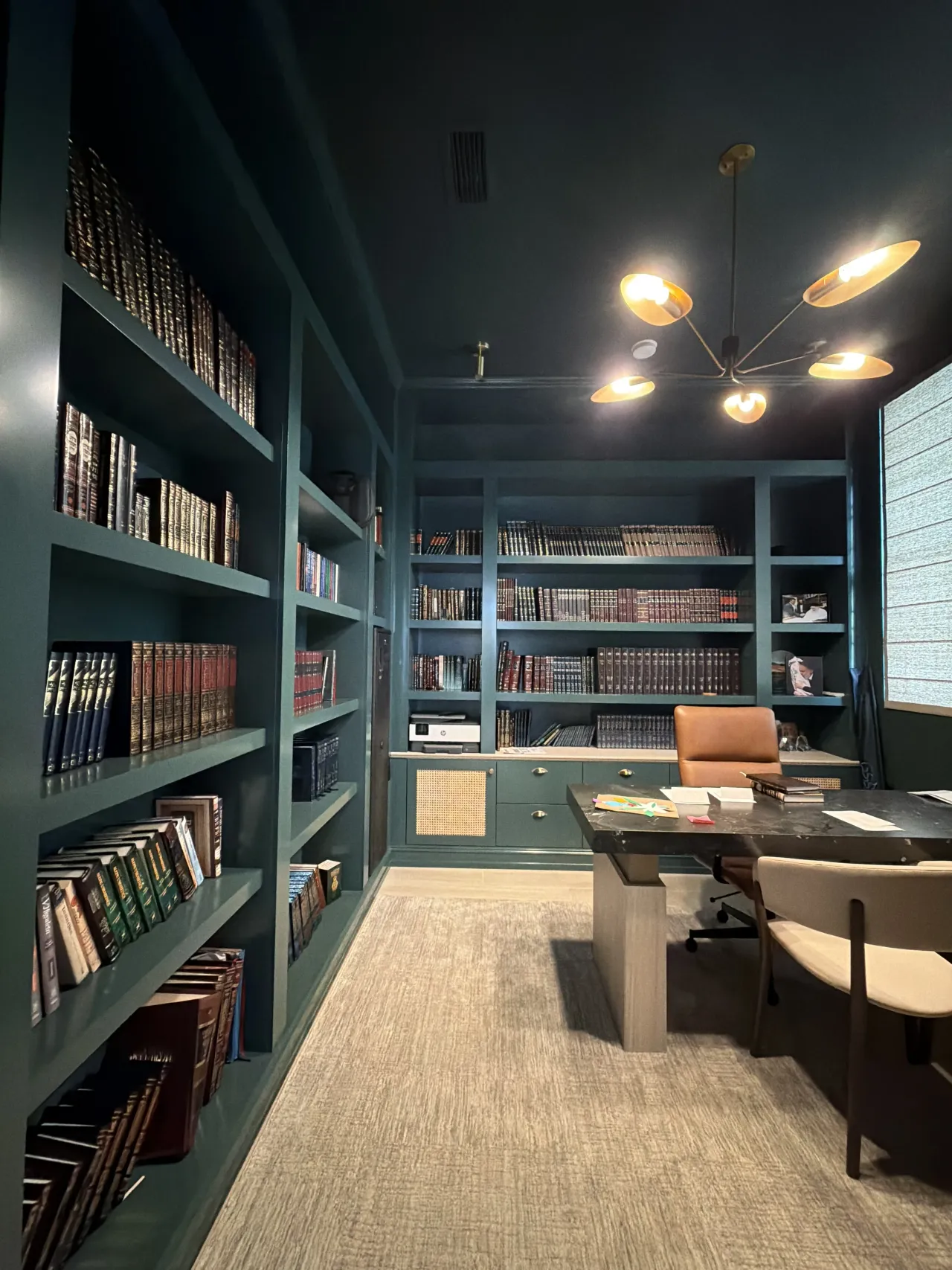 Dark green lacquered library wall with floor-to-ceiling bookshelves full of hardcover books, under a sputnik-style multi-arm chandelier, desk area
