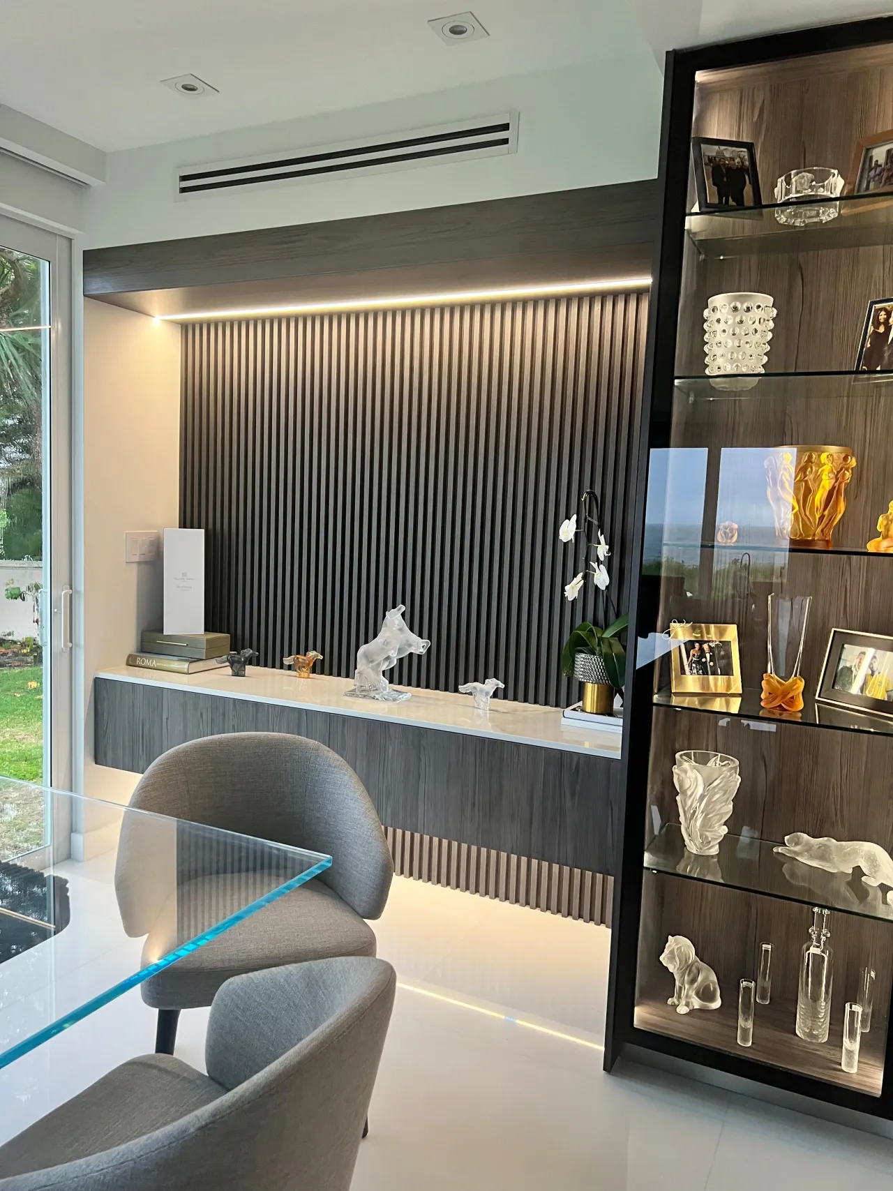 Dark slat accent wall behind a floating console with veined stone top holding glass sculptures, lit glass display cabinet at right, garden door at left