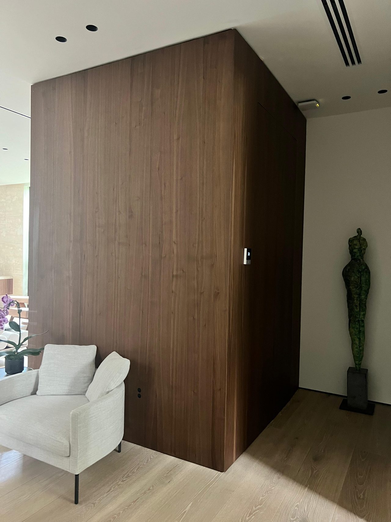 Large dark wood paneled cabinet/closet volume with a hidden door handle, beige armchair and sculpture nearby