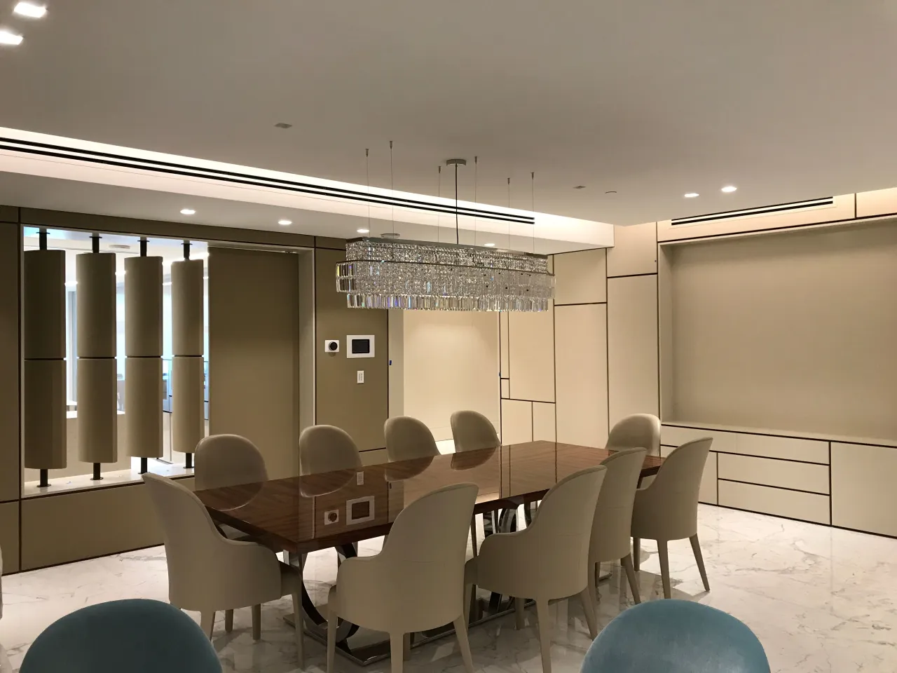 Dining room with a wood-grain table and upholstered chairs under a linear crystal chandelier, flush panelled wall cabinetry with a drawer bank at right
