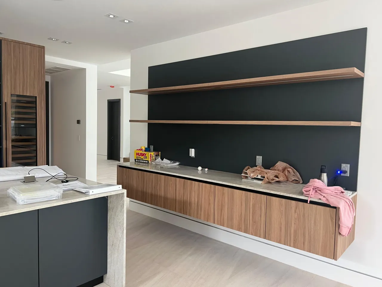 Floating wood-grain cabinet run with a light stone top and two long wood-grain shelves on a black accent wall, dark island with stone top at left