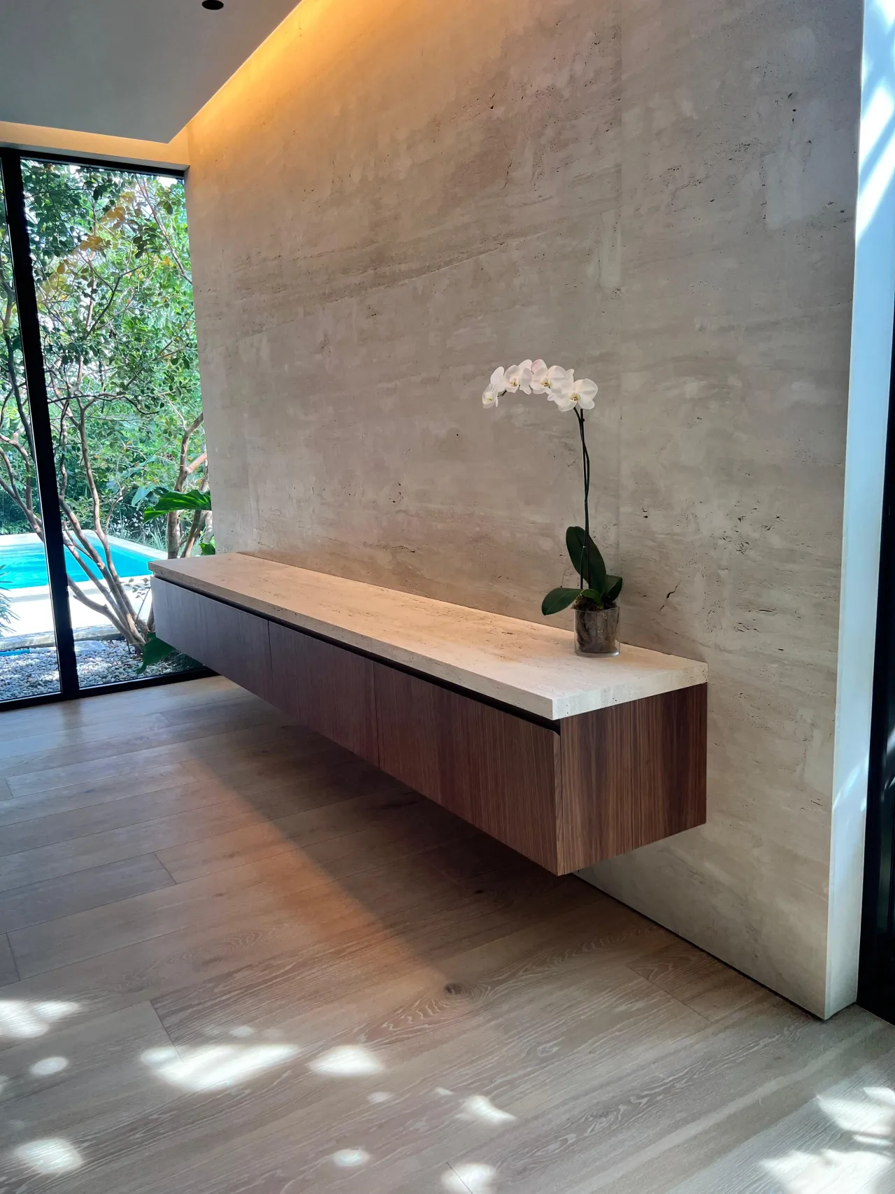 Long floating wood-grain console with a light stone top against a textured stone wall, single orchid on top, glazed wall showing a pool and trees