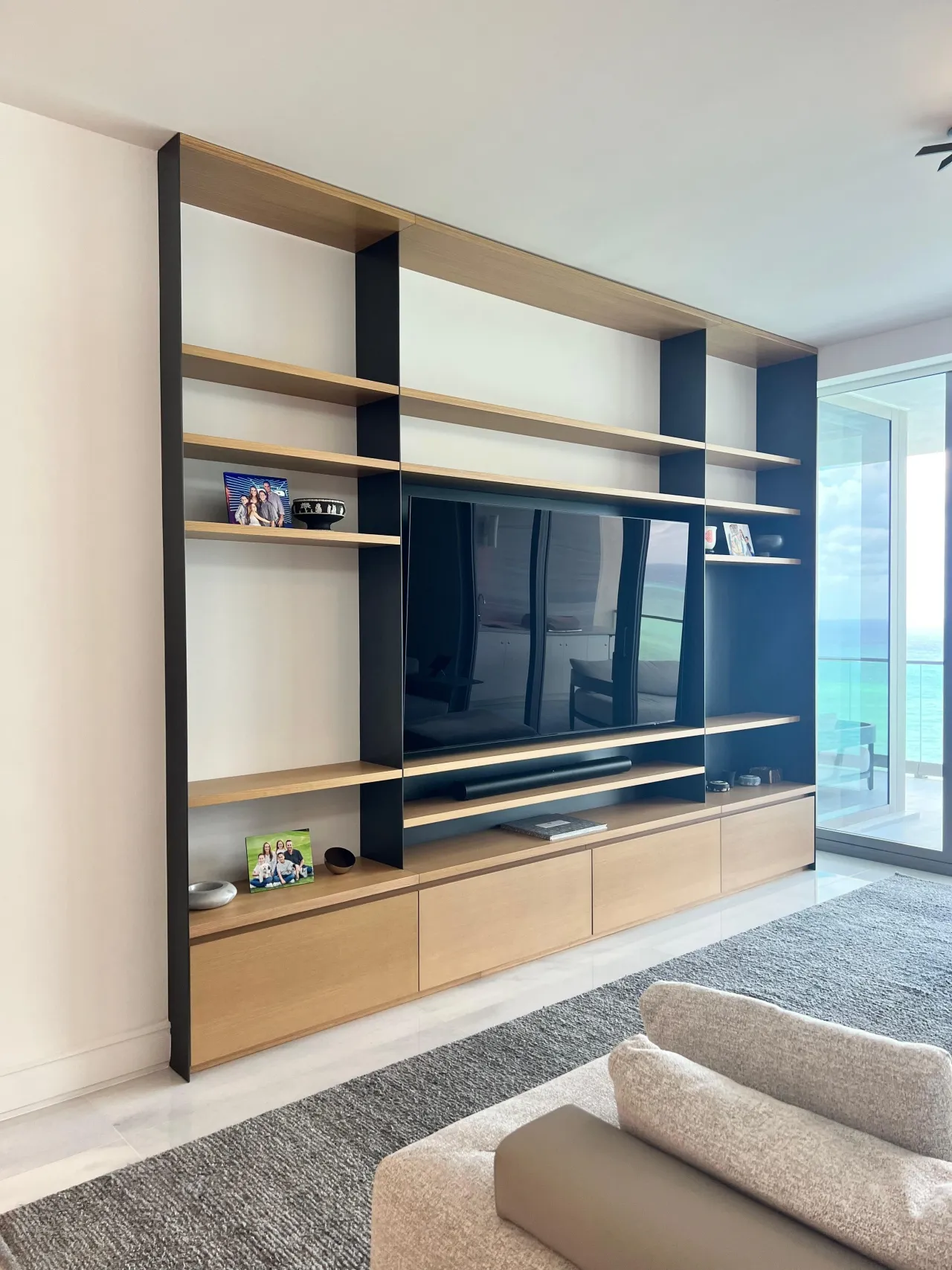 Floating wood-tone TV media wall with open shelving, framed family photos, and an ocean view through a glass balcony door