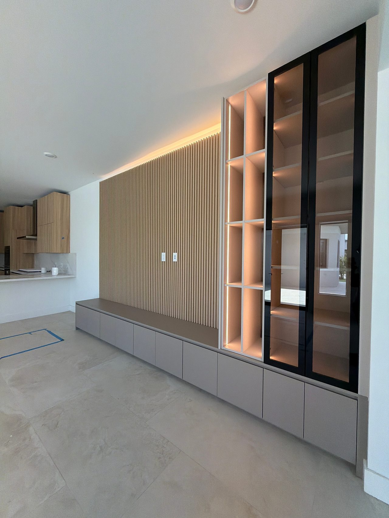 Floor-to-ceiling black-framed glass display cabinet beside a backlit white cubby column, wood-grain slat wall and low gray cabinets at left