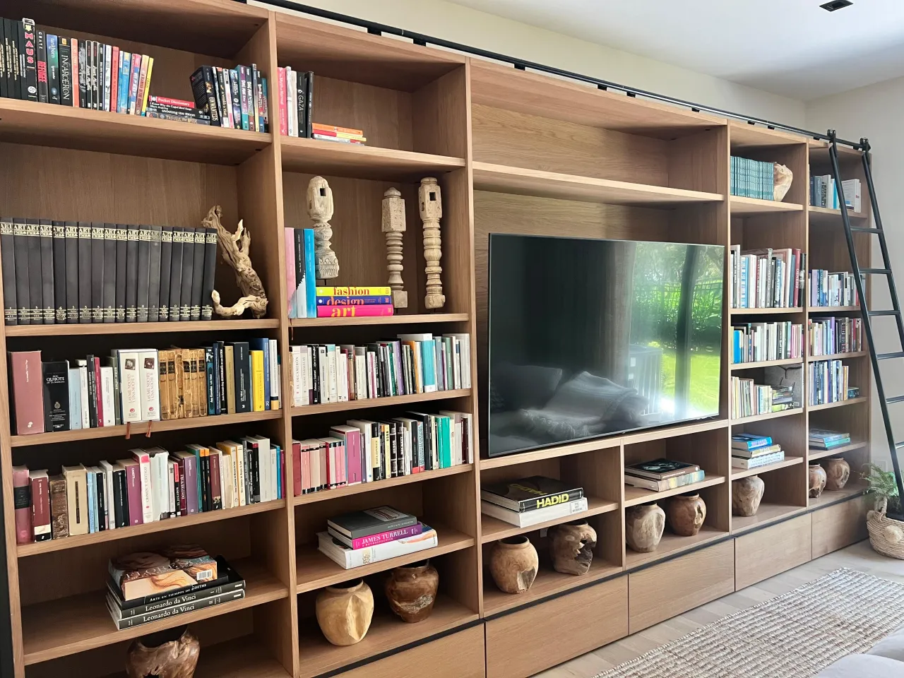 Floor-to-ceiling light wood bookshelf wall with a central TV niche, rolling library ladder, books, and decorative carved objects