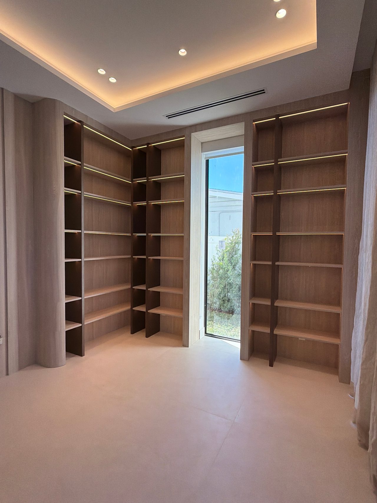 Room lined with floor-to-ceiling wood-grain open shelving with lit uprights, empty shelves, a cove-lit ceiling and a narrow full-height window