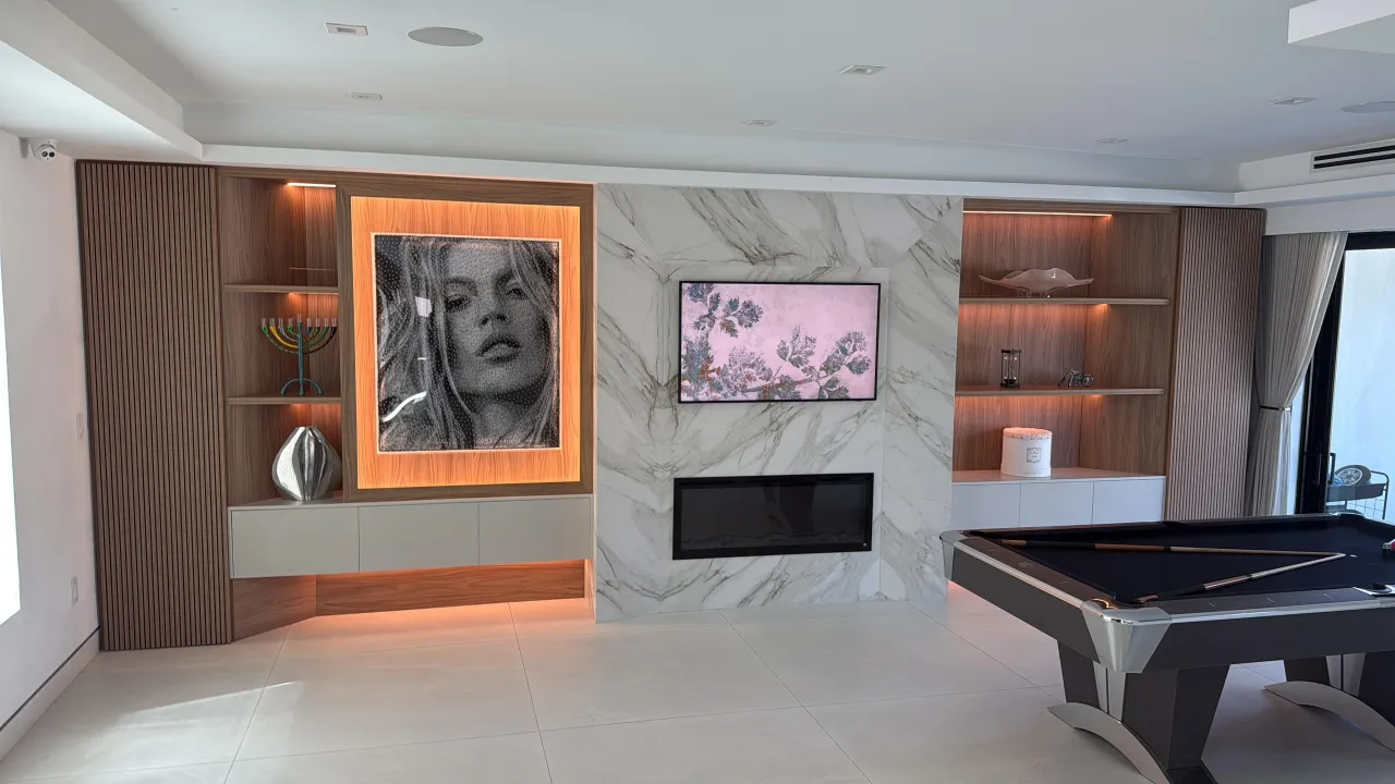 Game room with a veined stone wall holding a mounted TV above a linear fireplace, flanked by wood-grain shelving and a lit framed portrait