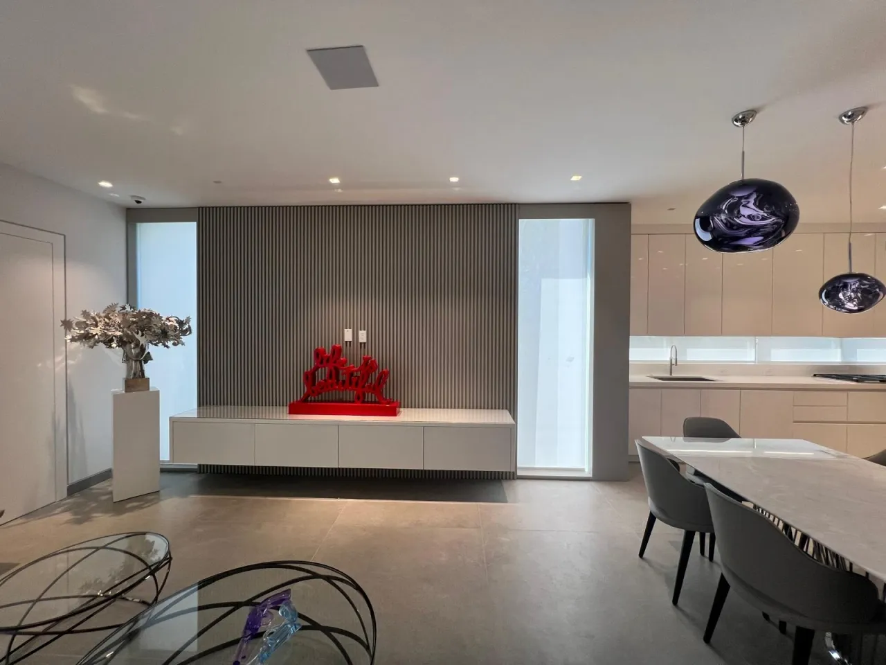 Gray fluted wall panel behind a low white console holding a red sculpture, with a white plinth of dried flowers at left and a kitchen beyond