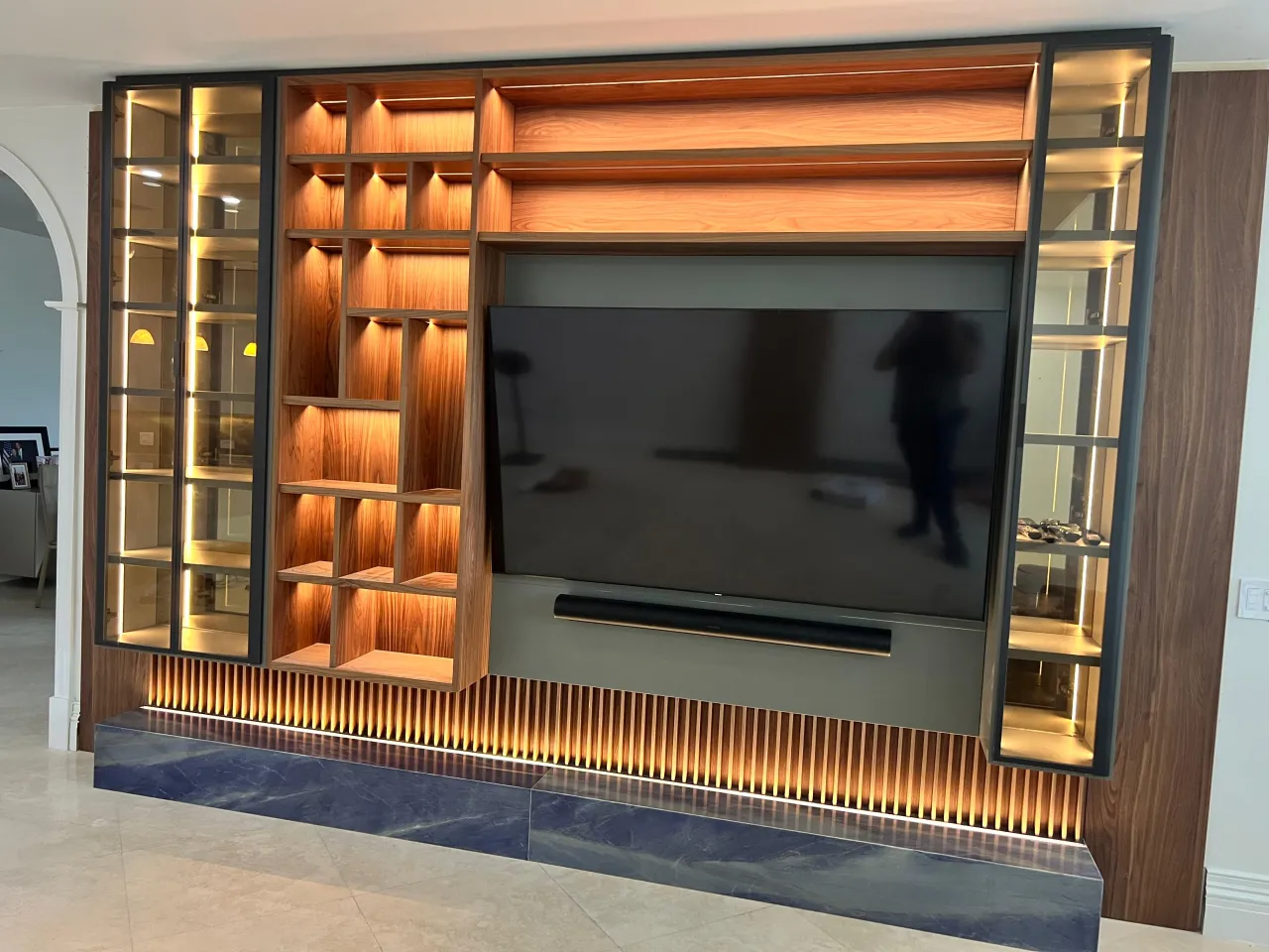 Illuminated wood-grain wall unit with lit open cubbies, glass cases each side, wall-mounted TV, and a backlit fluted strip above a dark stone base